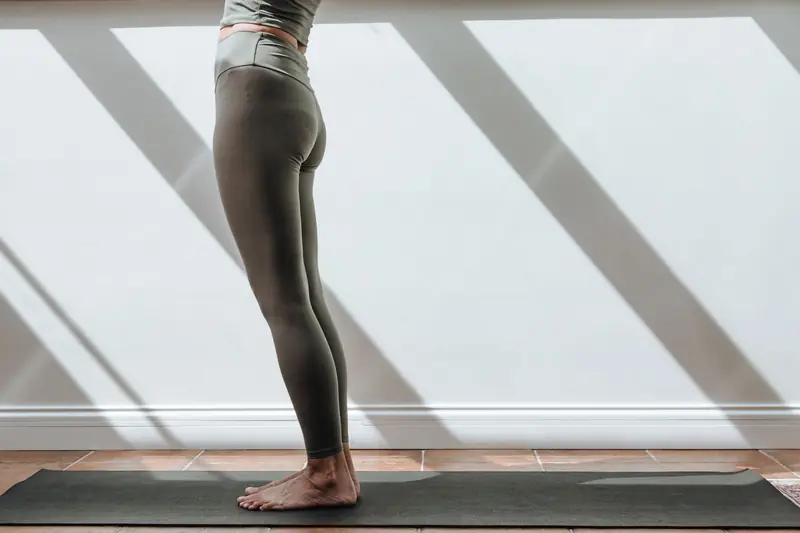 A woman standing barefoot on a yoga mat, wearing leggings in a sunlit room