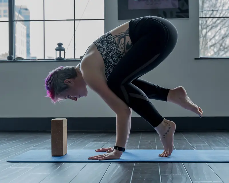 A woman performs a crow pose on a yoga mat indoors, focusing on balance and strength