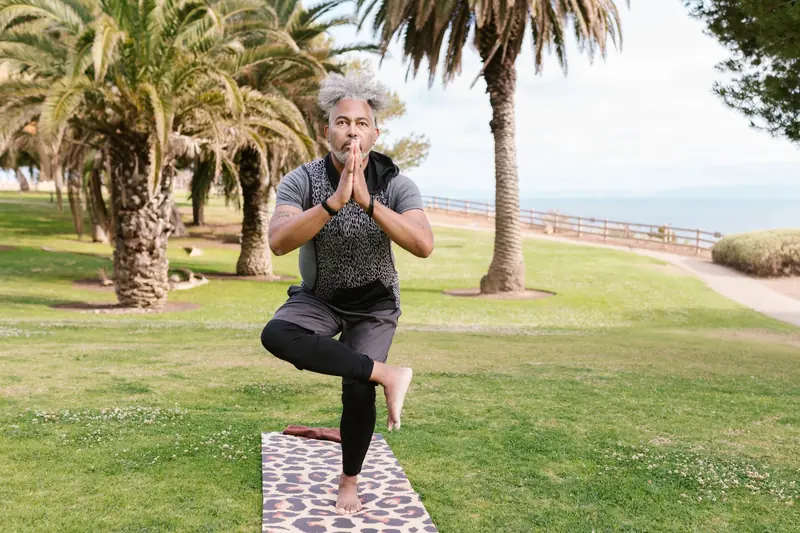 A man performs a tree pose on a yoga mat in a sunny park, surrounded by palm trees