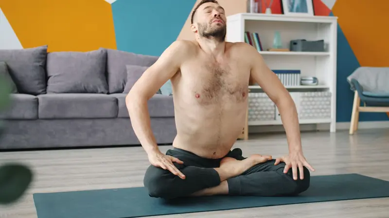 Shirtless man performing yoga meditation pose on mat in modern living room