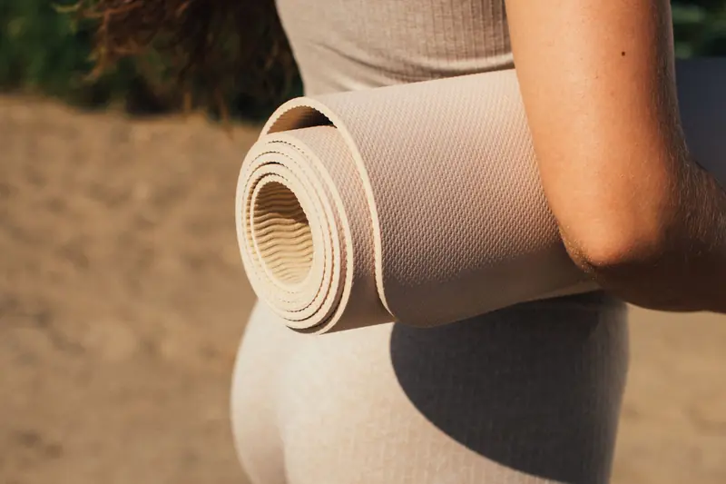 Person holding a rolled yoga mat in a sunlit outdoor setting, emphasizing fitness and wellness