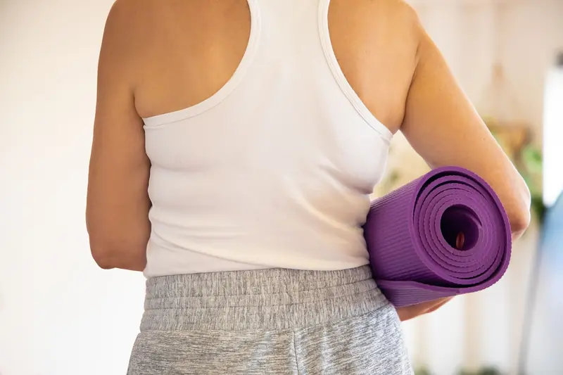 Woman in white tank top holding a rolled purple yoga mat indoors, preparing for exercise